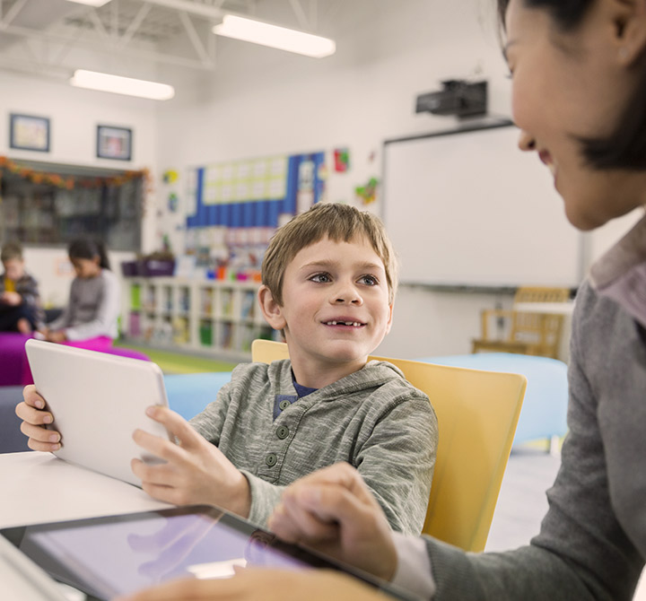 School teacher with young elementary student holding a tablet in a classroom.